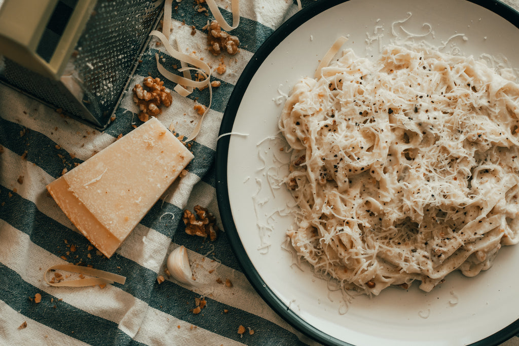 A plate of alfredo on a striped blue and white tablecloth with a wedge of parmesan and a grater 